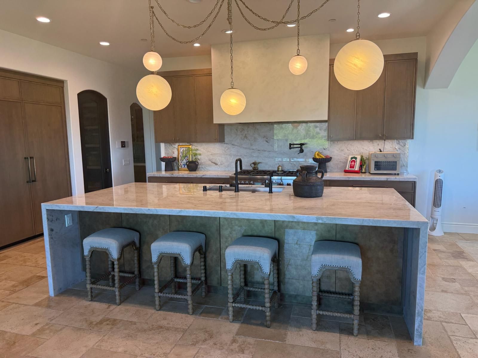 Remodeled Rancho Santa Fe kitchen with a waterfall stone island, four upholstered stools and globe pendant lights